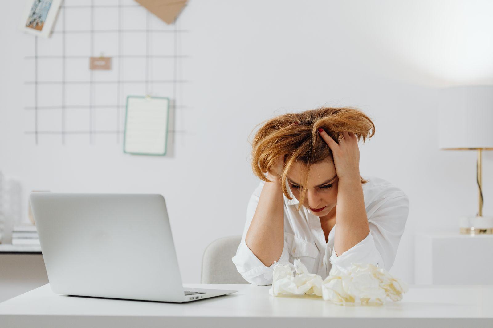 Femme visiblement débordée assise à son bureau, tête entre les mains devant un ordinateur portable et des papiers froissés