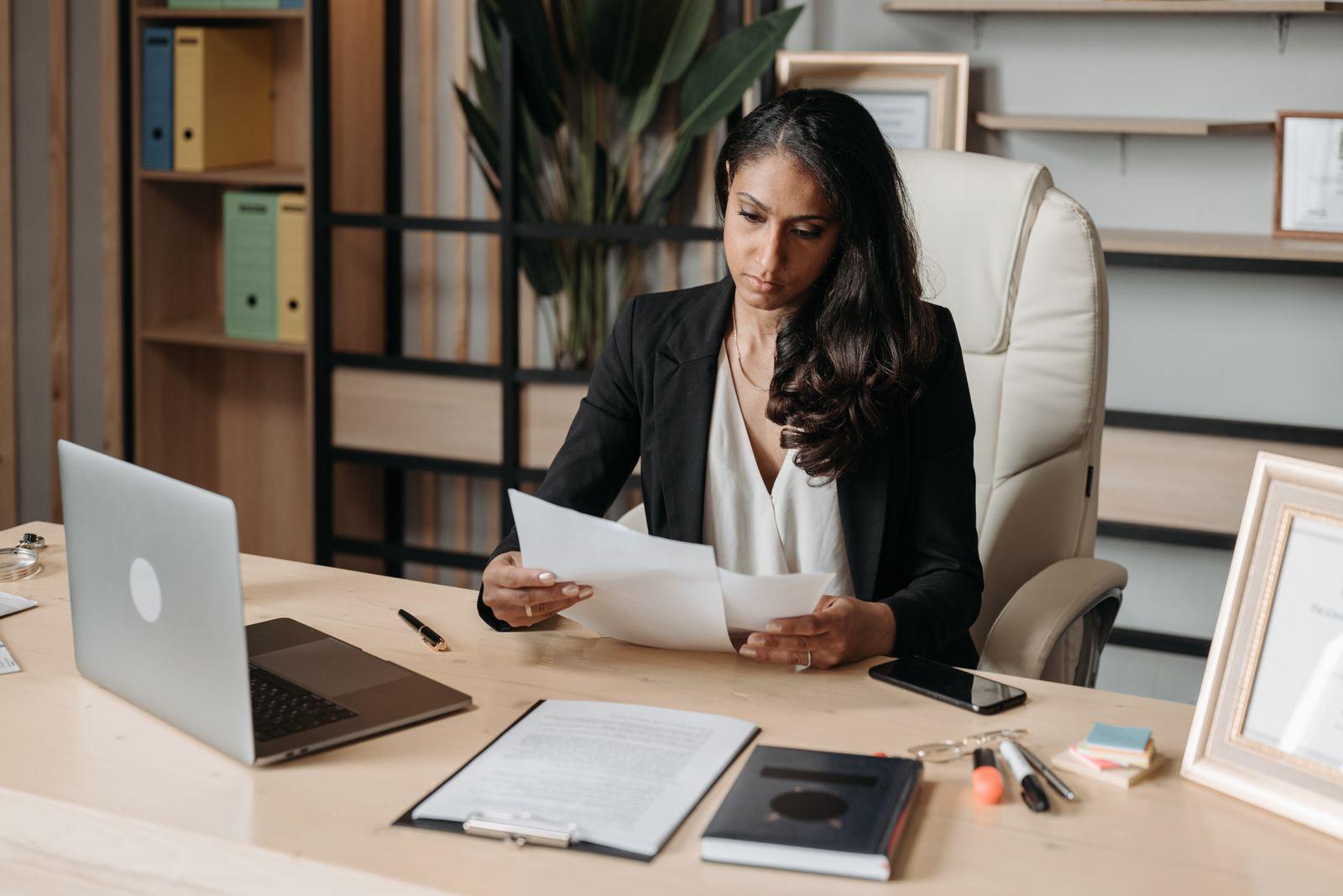Femme en tenue professionnelle debout à son bureau en train de consulter des documents, avec des classeurs rangés en arrière-plan