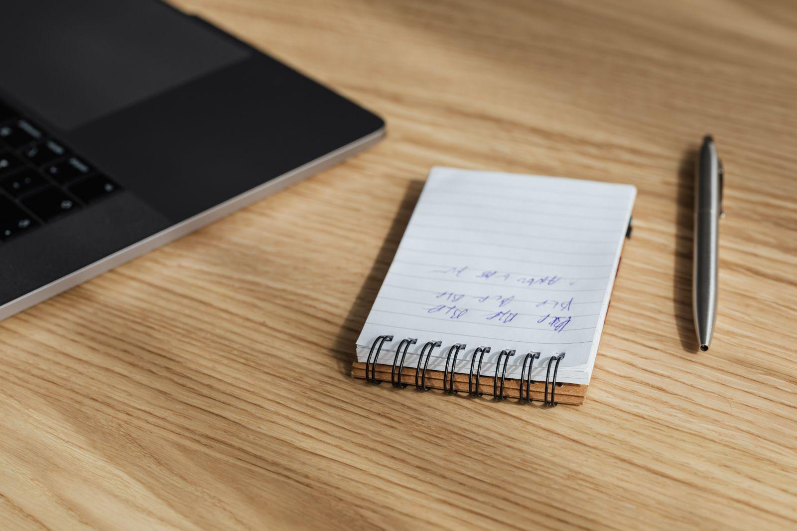 Carnet à spirale ouvert avec des notes manuscrites, posé sur un bureau en bois à côté d'un ordinateur portable et d'un stylo