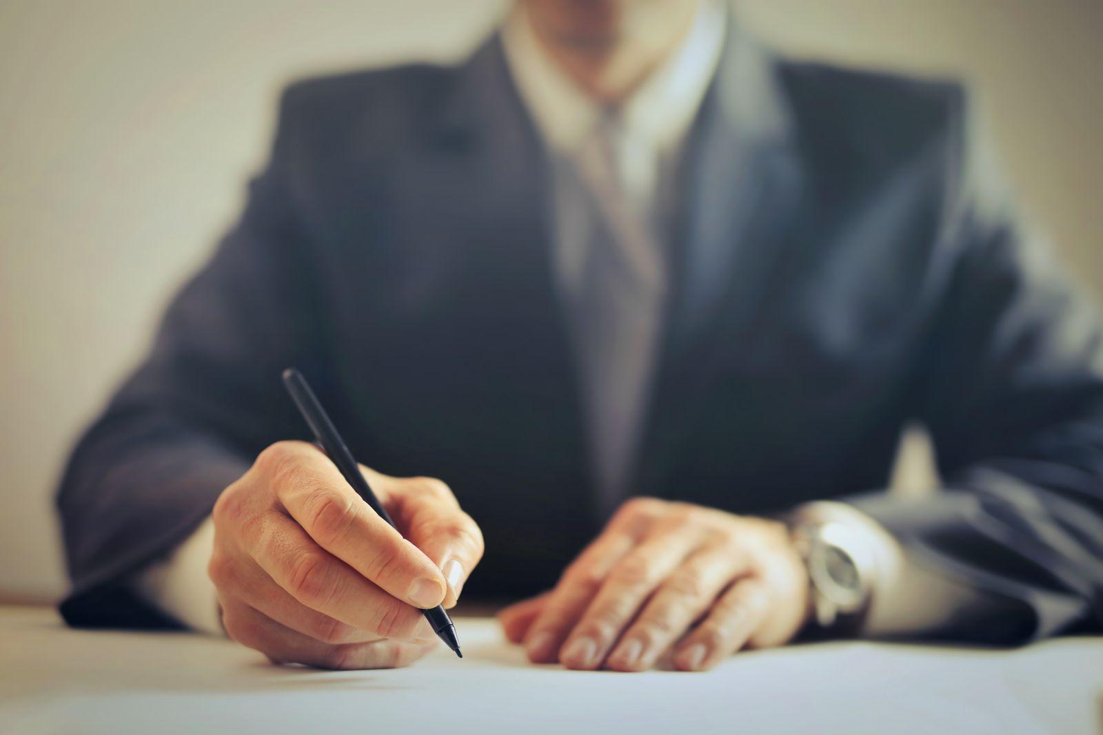 Homme en costume signant un document officiel avec un stylo, gros plan sur les mains et le document