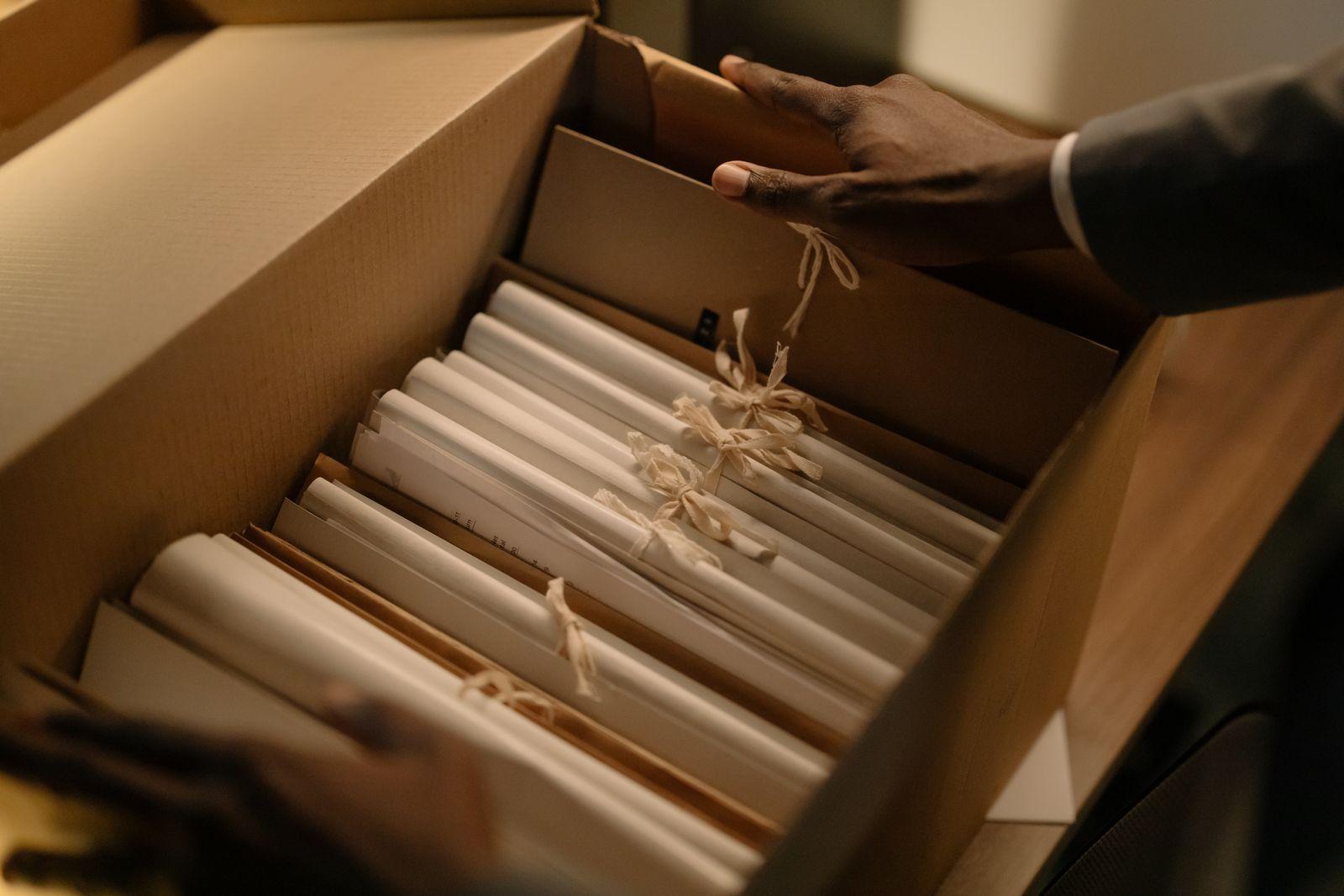 Mains rangeant des dossiers reliés par des liens dans une boîte en carton, évoquant un classement d'archives